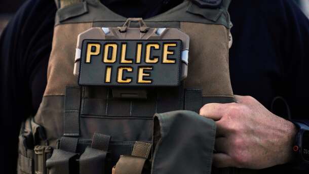 A light brown tactical vest is worn by an unseen man. One of his hands clasps the vest at the right side of the image. In the chest of the vest, a badge reads "Police, ICE."