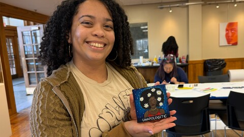 A Black woman smiles and holds up a small square painting of a woman with flowers in her hair. The painting reads "unapologetic."