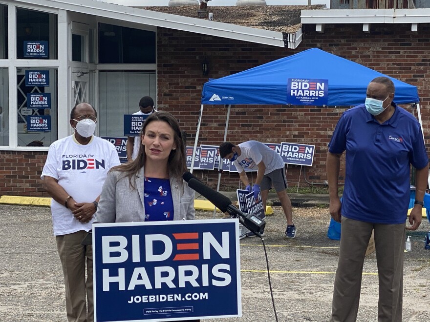 Woman in blue blouse and grey blazer speaks at a podium with a sign in front of her that reads "Biden Harris"