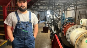 A man wearing overalls stands at the foot of a staircase, and to his right are large pipes. 