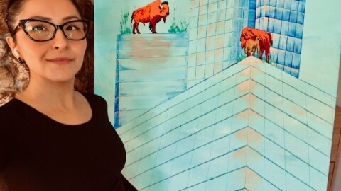 A young woman with dark glasses and a black long-sleeve shirt stands next to a big square painting. In the painting, a beaded male bison stands on the top of one tall building, while a beaded mama and baby bison cuddle on the top of another tall building.