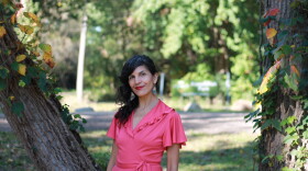 image shows an Indigenous woman dressed in a rose-pink dress. she is wearing lipstick and her hair is tied into a side ponytail. she poses in between two cottonwood trees.