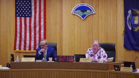 A man in a blue suit and yellow tie and a woman in a white and purple blouse sit on the left and right, respectively, of a long desk with mics and name plates to denote their positions as county commissioners. An American flag hangs on the wall on the left, while a Michigan flag hangs on the right. 
