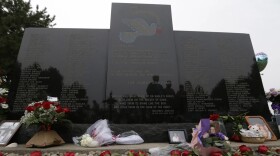 A wide, dark memorial stone takes up the frame. Red roses and pictures line the bottom of the stone. 