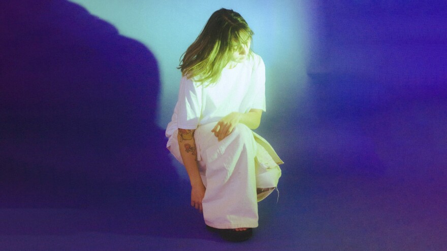 A woman in white, loose-fitting clothing crouches in an empty space with blue coloring all around her.