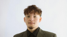 A man in a green button blazer and a black undershirt stands in front of all white background 