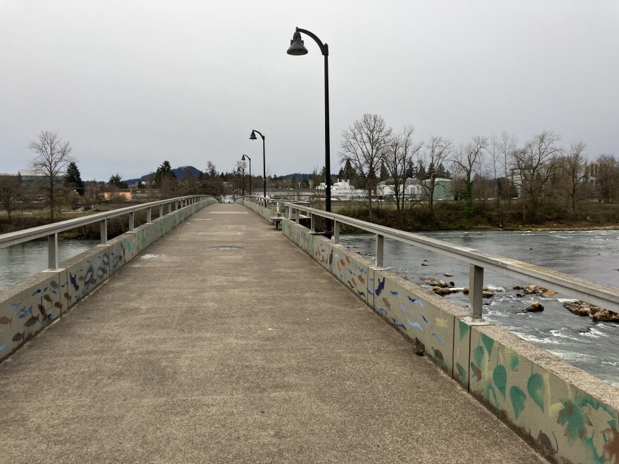 A bike/pedestrian bridge over the Willamette River. Lamp posts line the bridge. Trees and buildings are on the far riverbank. 