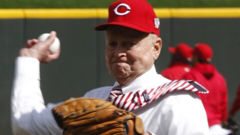 a man in a tie, white button-up shirt, black pants and a cincinnati reds ball cap throws a ball from a pitcher's mound in a baseball field