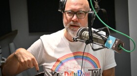 A man inside a radio studio gestures with his right hand while talking at a microphone. He is wearing a T-shirt that reads "KC Rainbow Tour."
