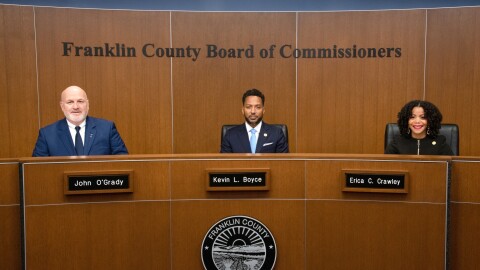 A white man, a Black man, and a Black woman sit at a large, curved desk with Franklin County's seal on it. Their name plates read, from left: John O'Grady, Kevin L. Boyce, and Erica C. Crawley. 