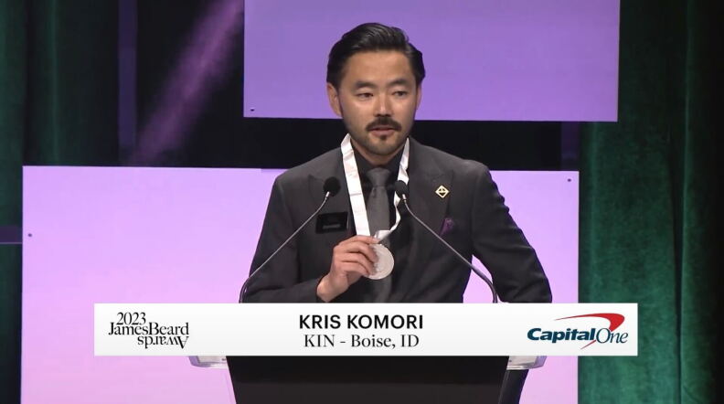  A man stands in front of a podium with two small microphones holding a medal. There is a lower third that says 2023 James Beard Awards, Kris Komori KIN - Boise Idaho