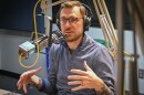 A man sitting inside a radio studio talks at a microphone and gestures with both hands.