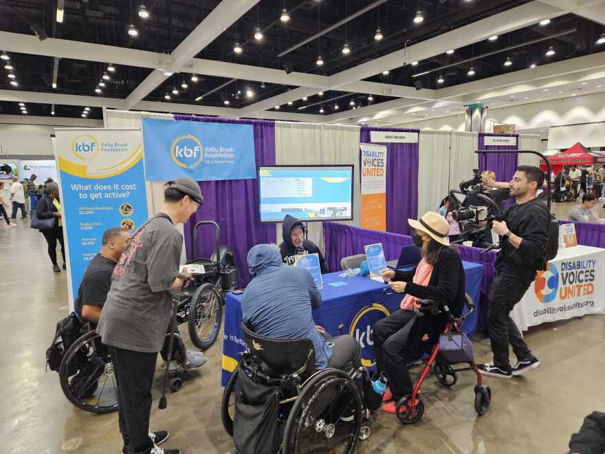 A photo of people, some standing, some sitting in wheelchairs, gathered around an expo booth with light blue signage with the logo "Kelly Brush Foundation"