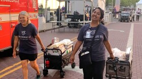 Two woman wearing matching t-shirts promoting food rescue pull cloth wagons filled with containers of food down a brick road. 