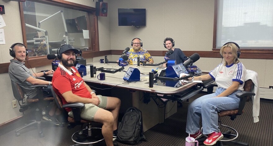 Five smiling people wearing headphones sit at a table in a radio talk studio; the man on the left in the foreground has short dark hair and is wearing a grey short sleeved polo shirt, grey shorts and sneakers; the man on the right in the foreground has a dark beard and is wearing a black baseball cap, a red short sleeved polo shirt with a white collar, white writing, a yellow logo and black stripes on the shoulders and khaki shorts; the man on the left in the background is bald with a blonde beard and is wearing glasses and a yellow, blue and white patterned soccer jersey; the man on the right in the background has short dark hair and is wearing a blue long sleeved shirt; the young woman at the end of the table has long blonde hair and is wearing a white soccer jersey with blue stripes on the shoulders and a red logo.