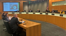 Two women,and a man sit at a long wooden table, with microphones on the table. They are addressing the Normal Town Council in the council chambers.