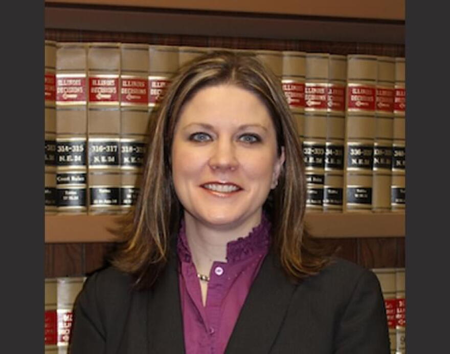 A woman in a dark blazer and purple blouse standing in front of a bookshelf filled with law books.