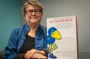 A woman sits at a table with her hands folded in front of her. Beside her, on the table, stands a large, coffee-table book called "The Jayhawk, the Story of the University of Kansas's Beloved Mascot."