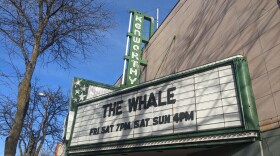 A white marquis spells out THE WHALE in black lettering on the front of an old theater with a green sign that reads "Kenworthy" against a blue sky.