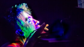 A man is squatting while starring into a screen, while red blue and green lights shine brightly into his face and neck.
