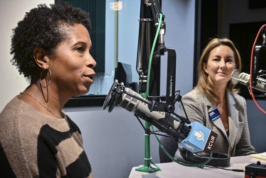 Two women sit inside a radio studio. They are seated at microphones. One on left (Brandy Williams) is talking while the other (Julie Donelon) is listening.