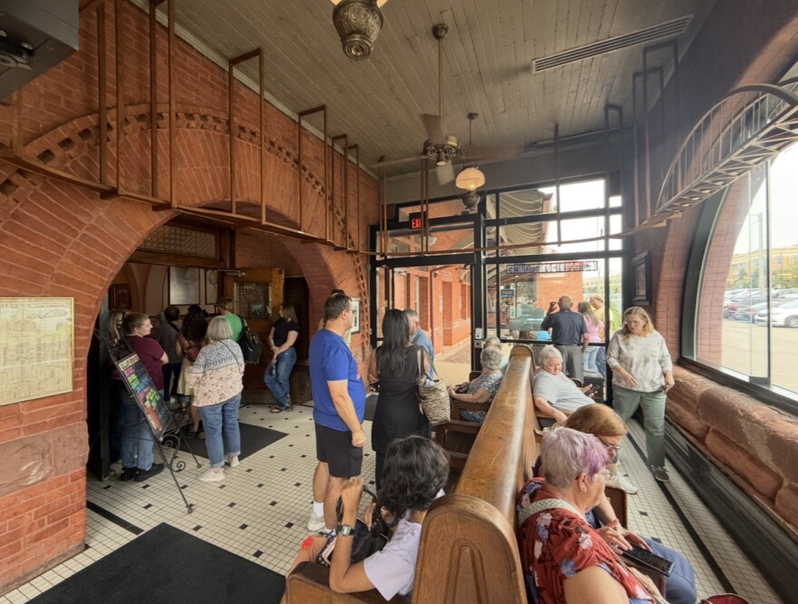 A crowd of people is seen standing in line and sitting on benches in a waiting room of a restaurant. The floor has vintage tiling, and the room is surrounded by large windows. 