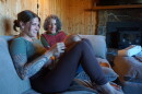  A woman with tattoos on her right arm holds a small baby in her lap on a grey couch. Another woman sits next to her, gazing at the baby.