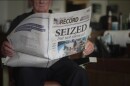 An older man sits in a desk chair and holds the Marion County Record newspaper published after the outlet was raided by local police. The headline on the front page, in large text, reads "SEIZED...but not silenced." There's a story on the right side of the front page that has the headline "KBI takes over," with a one-column line of text. Next to that text is a four-column image of the police raiding the newspaper office. On the back page of the paper is a single, wide column of text, with a large quote above it reading, "Freedom of the press is as important to democracy, as democracy."