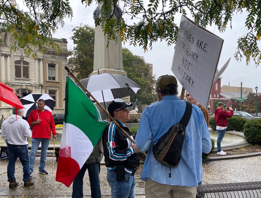 A man with his back to the camera carries a sign, another man talking to him holds an Italian flag