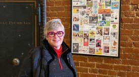 Woman wearing red glasses and a dark coat standing in front of a security safe and a brick wall with a collage of advertisements hanging in a display frame