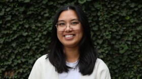 a woman with black hair, wearing glasses and a cream colored cardigan stands in front of a green ivy wall