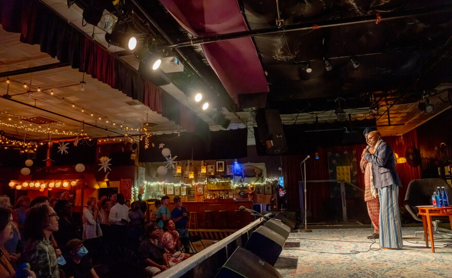 A large audience sits inside a music venue watching two speakers onstage.