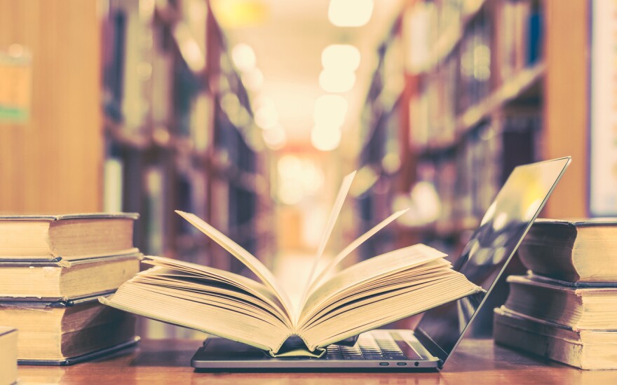 Two stacks of books frame an open laptop, with an open book and its pages sitting atop the keys. The tall stacks of library bookshelves are blurry in the distance.