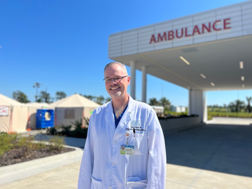 A doctor stands outside the ambulance bay of the hospital he works at. In the background medical tents are set up.