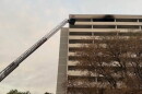 Fire trucks with extended ladder respond to a fire on the upper floor of a multi-story building, with black smoke visible from the windows. Trees and emergency lights are also visible in the scene.