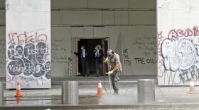 In this July 8, 2020 photo, a worker washes graffiti off the sidewalk in front of the Mark O. Hatfield Federal Courthouse in downtown Portland, as two agents with the U.S. Marshals Service emerge from the boarded-up main entrance to examine the damage. Protests and clashes with city, state, and, more recently, federal law enforcement have become a nightly occurrence in Oregon’s largest city. CREDIT: Gillian Flaccus/AP