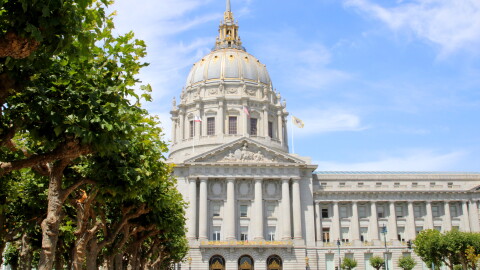 A view of the front of city hall with a few trees in the foreground and a blue sky with wispy clouds behind it. The city hall building is long and rectangular with columns in front, gold trim, and a big dome coming out of the center of it. 