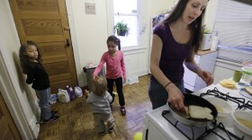 A woman with long brown hair is making a grilled cheese sandwich while her three children play behind her in the kitchen. 