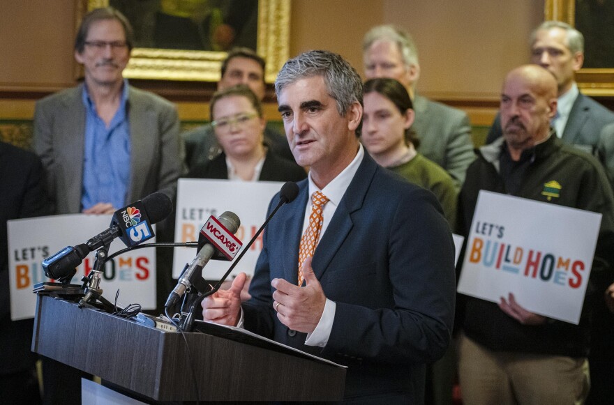 A man wearing a suit stands at a podium indoors; other people stand behind him holding signs that say "let's build homes"