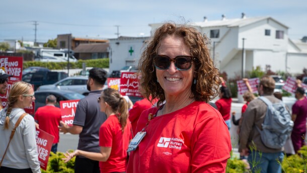 A woman with curly brown hair wearing a National Nurses United t-shirt smiles at the camera. Behind her, people hold up signs on a street corner.