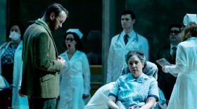 Dr. Oliver Sacks, played by Jarrett Porter, is dressed in a brown suit coat and looks to Susanna Phillips, who sings the role of the patient Rose in the opera “Awakenings." Phillips is wearing a hospital gown and is looking distantly while in a wheelchair.