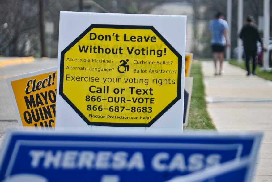 A white sign with a yellow octagon printed on it reads "Don't Leave Without Voting." Two people are walking in the background and there are other political campaign signs in front and behind it.