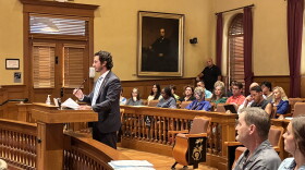 A man in a suit speaks at a podium in a courtroom filled with seated people. A large portrait hangs on the wall behind him. Papers are on the podium and benches.