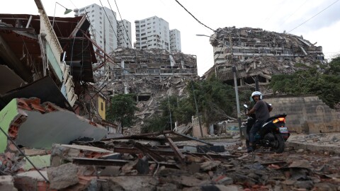 A man in a bike rides through a street surrounded by collapsed buildings. 