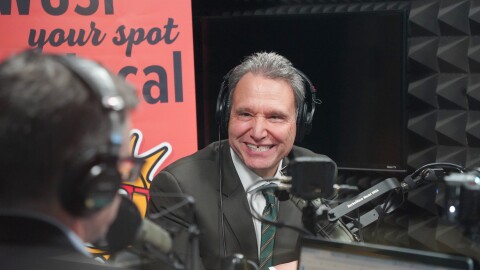 Man in dark green suit and tie smiles as he sits by a microphone in a studio 