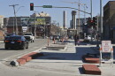 Exterior photo of a busy street at left. At right two people are walking away from the camera on a freshly poured concrete space.