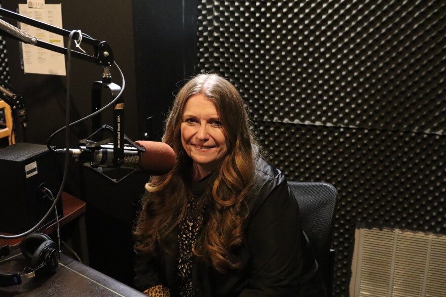 A woman sits smiling in a soundproofed radio studio beside a mounted microphone.