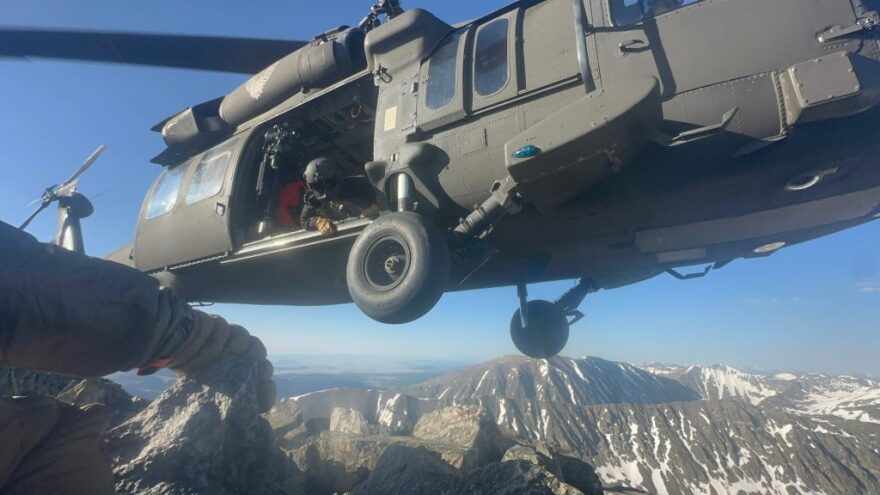 A person wearing a helmet leans out of a helicopter above snowcapped peaks. 