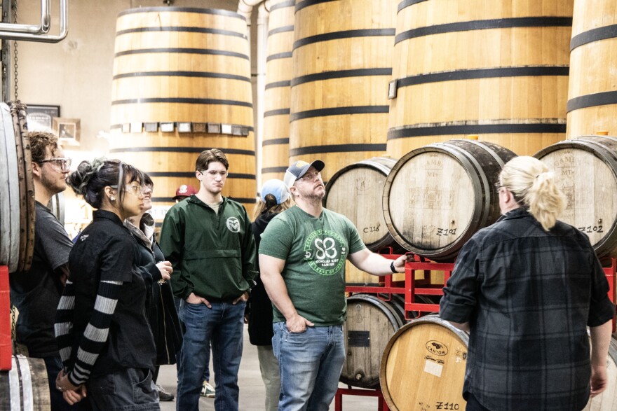 A few students wearing safety glasses are standing in a group - a young man with brown hair, a young woman with black hair wearing a black tee shirt and a grey and black-striped long sleeve underneath it, a young man with a dark green windbreaker and jeans, and a man wearing a green and cream ballcap, a green tee shirt and jeans. Behind them are very tall and wide wooden barrels, and to the right are some smaller wooden barrels on their sides. To the right is a woman with blonde hair wearing a checkered long-sleeve facing the group.