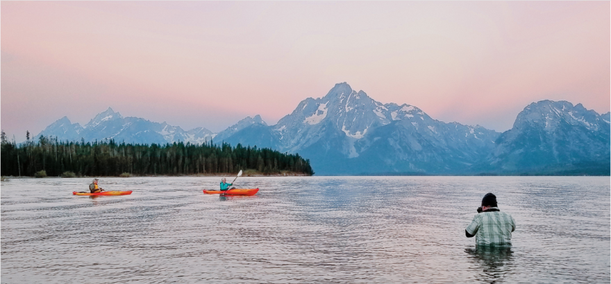 Two people kayaking on a calm lake with a mountainous backdrop at sunset. The sky is pink and purple. A person in the foreground is standing in the water, taking a photograph.